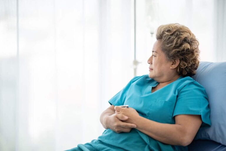 A woman in medical scrubs sits on a hospital bed, looking out a window with her hands clasped in her lap.