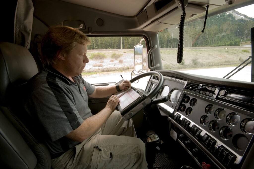 A man sits in the driver’s seat of a semi-truck cab, filling out paperwork on a clipboard, with various controls and gauges visible on the dashboard.