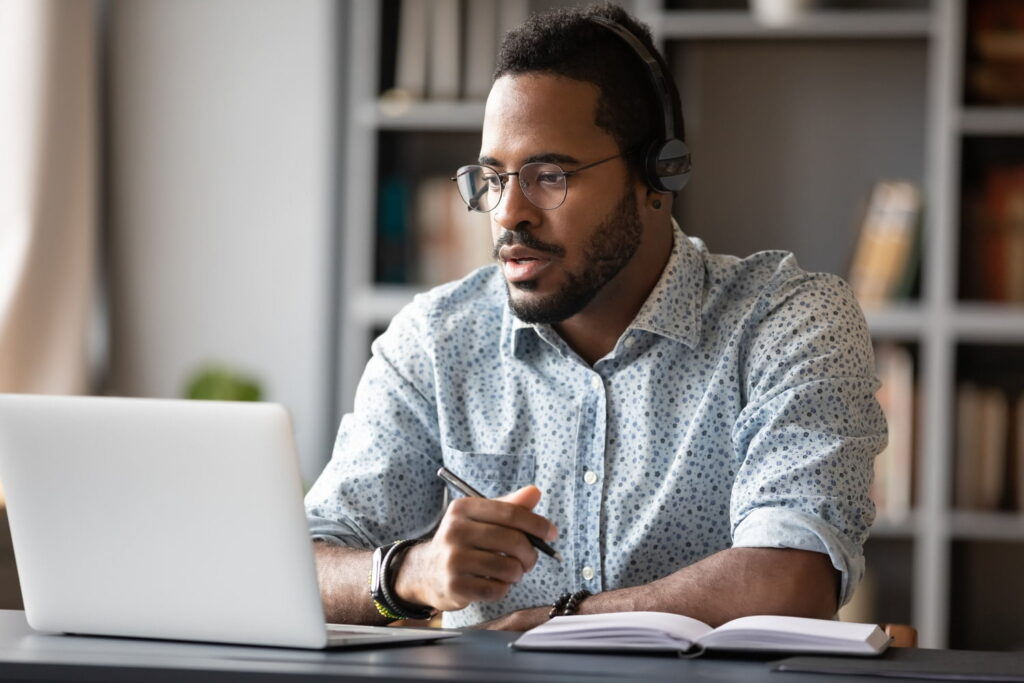 Man wearing headphones and glasses sits at a desk with a laptop and notebook, appearing focused while participating in an online meeting or class.