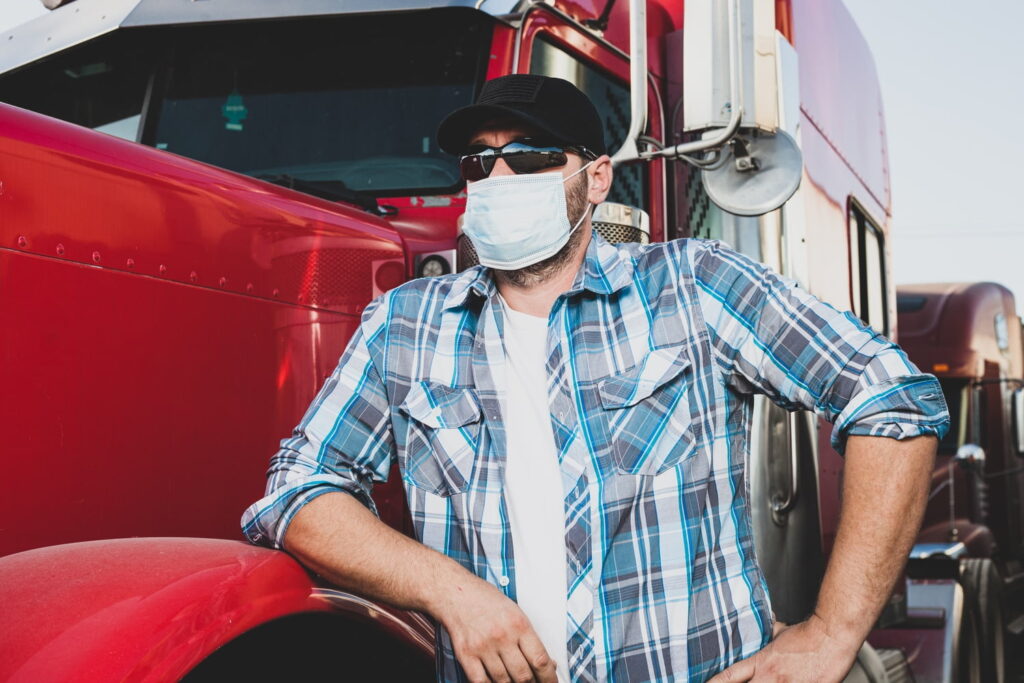 A man wearing a face mask, sunglasses, and a plaid shirt stands with his hand on his hip next to a red truck.