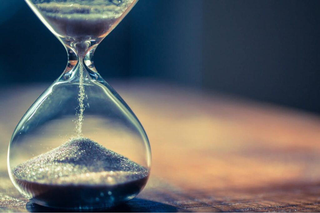 A close-up of an hourglass with sand flowing from the upper to the lower chamber, placed on a wooden surface with a blurred background.