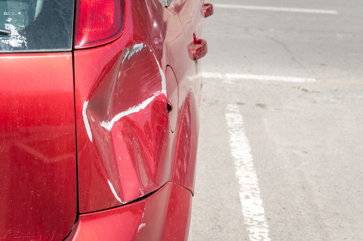 A red car with a large dent and scratches on the left rear side, parked in a lot with visible white lines on the pavement.
