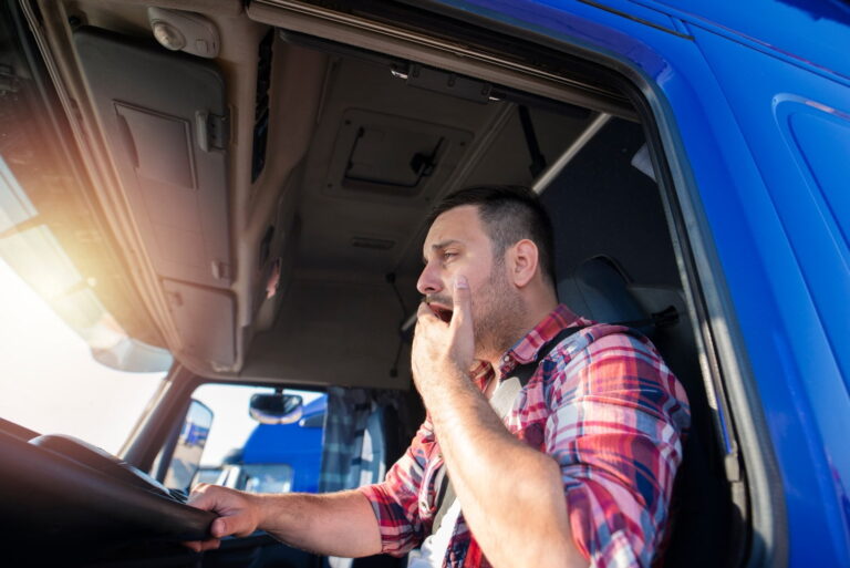 A man in a plaid shirt sits in the driver’s seat of a blue truck, yawning and appearing tired, with one hand on the steering wheel and the other covering his mouth.