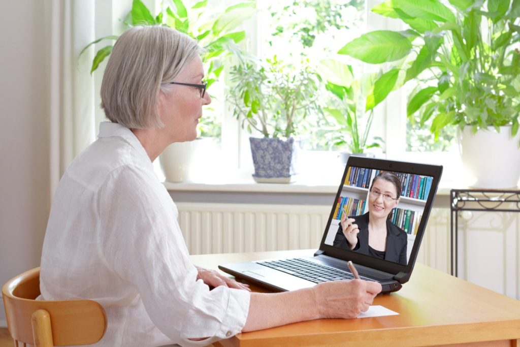 An older woman sits at a table, taking notes while having a video call with another woman on her laptop; both appear engaged in conversation.