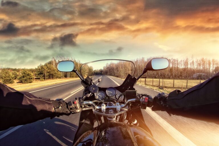 View from a motorcycle rider’s perspective on an empty road at sunset, with hands on handlebars and trees lining both sides of the road.