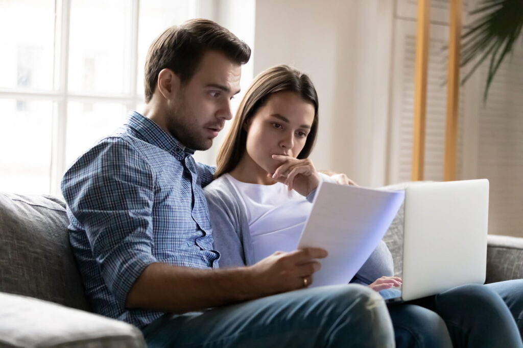 A man and woman sit on a couch looking at papers and a laptop screen with concerned expressions.