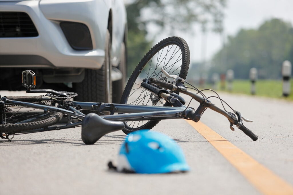 A bicycle lies on its side near the front of a car on a road, with a blue helmet on the ground in front.