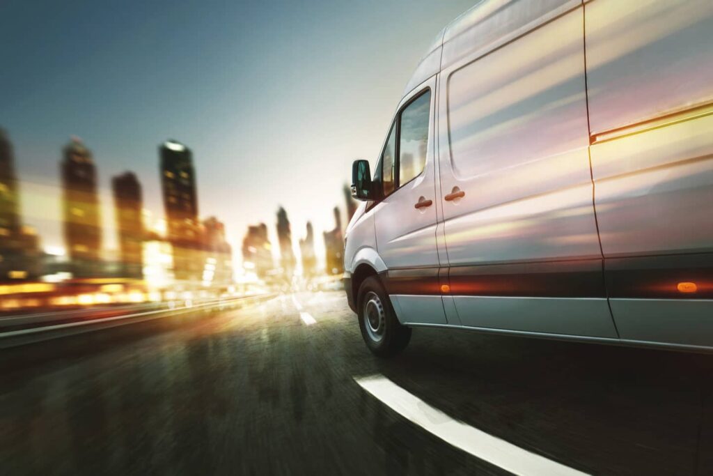 A white delivery van drives on an urban road at dusk, with blurred city lights and buildings in the background, suggesting motion and speed.
