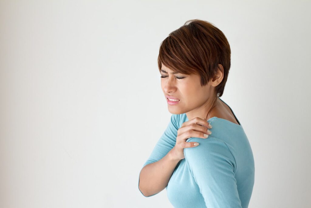 Woman in a blue shirt holds her left shoulder and appears to be in pain, standing against a plain white background.