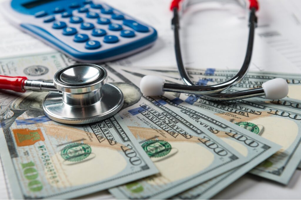 A stethoscope and a calculator rest on top of several US hundred-dollar bills, symbolizing healthcare costs and financial calculations.
