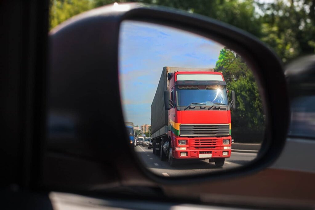 A red semi-truck is visible in the side mirror of a car, driving on a road lined with trees under a blue sky.