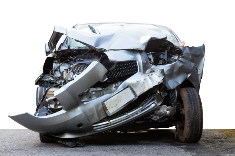 A severely damaged car with a crumpled front end is shown on a gray surface, isolated on a white background.