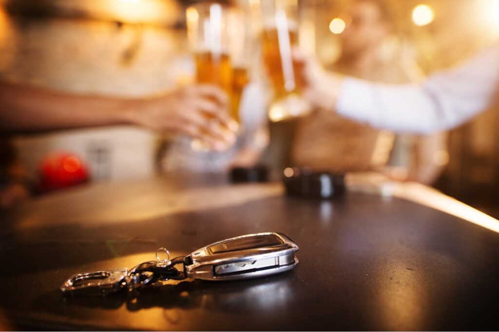 A set of car keys rests on a table in the foreground while people hold up glasses of beer together in the blurred background.