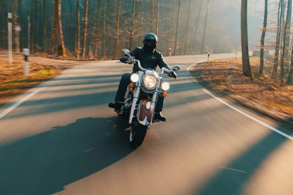 A person wearing a helmet and dark clothing rides a motorcycle on a winding road through a sunlit forest.