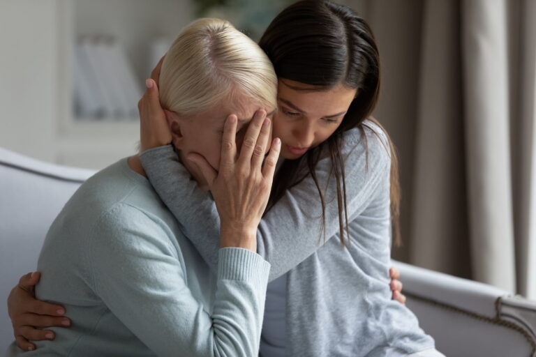 A young woman hugs and comforts an older woman who is covering her face with her hand, both seated indoors.