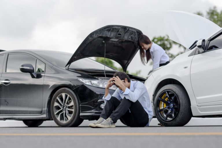 Two cars with open hoods are stopped on the road. A man sits on the ground with his head in his hands, while a woman checks under the hood of one car.