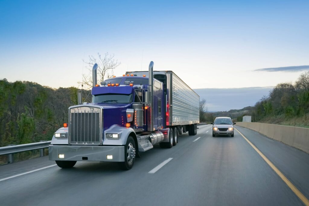 A purple semi-truck and a car drive side by side on a highway bordered by trees and a concrete barrier under a clear sky.