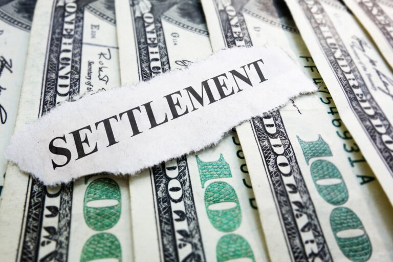 A torn piece of paper with the word "SETTLEMENT" sits on top of several spread out U.S. one hundred dollar bills.