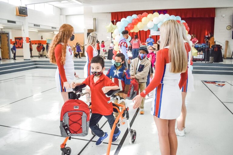 Children, some with mobility aids, enter a gym decorated with balloons, greeted by women in matching red and white uniforms.