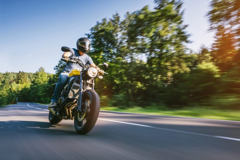 A person wearing a helmet rides a motorcycle at speed on a curved, tree-lined road under a clear sky.