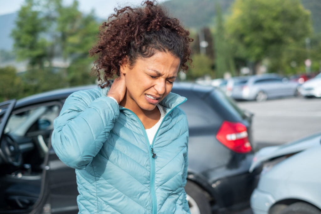 Woman holding her neck in pain stands near two cars involved in a rear-end collision in a parking lot during the day.