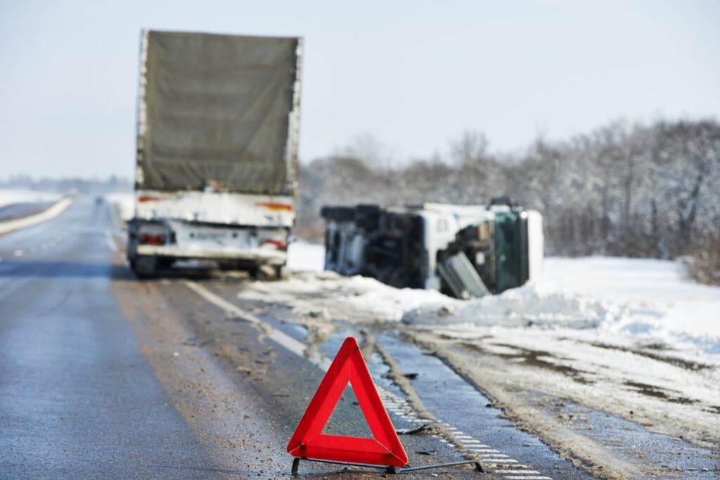 A truck lies on its side off a snowy road while another truck is parked nearby; a red warning triangle is placed on the roadside.