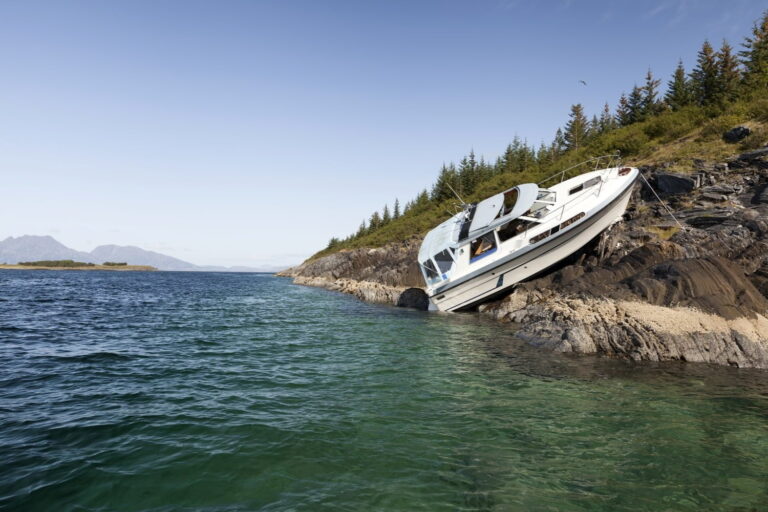 A white boat is stranded on rocky shore, tilted to one side, with green trees and calm water in the background under a clear sky.