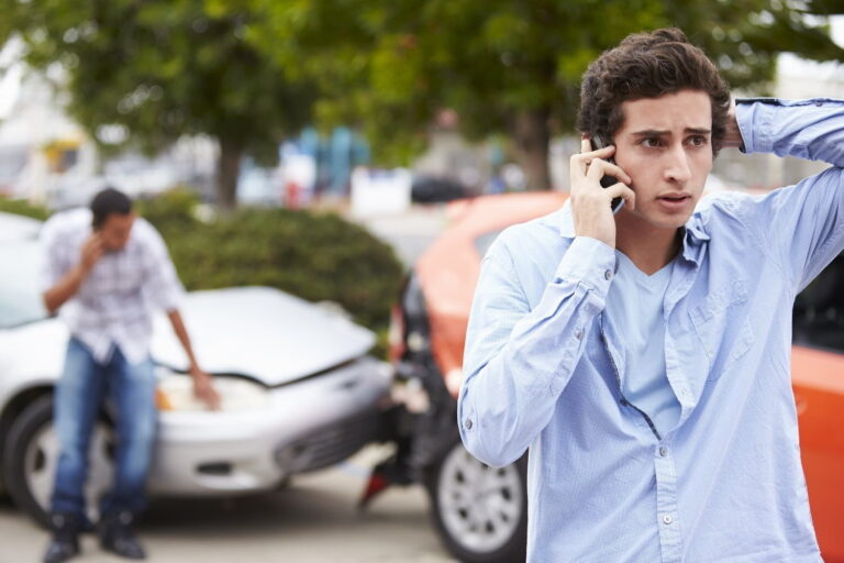 Two men are on their phones near crashed cars; one man stands in the foreground looking concerned, while another stands by a damaged white car in the background.