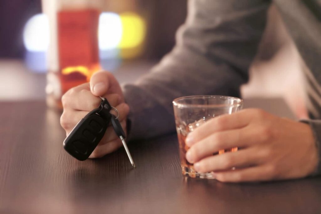 A person holds a car key in one hand and a glass of alcohol in the other while sitting at a table.