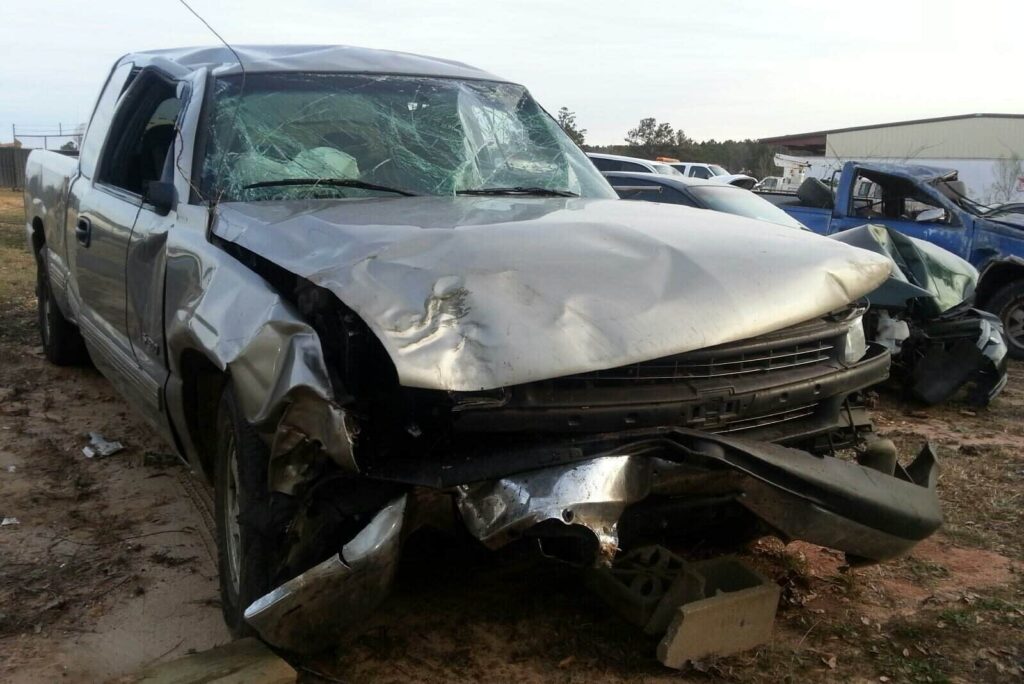 A heavily damaged silver pickup truck with a smashed front end and shattered windshield, parked in a junkyard alongside other wrecked vehicles.