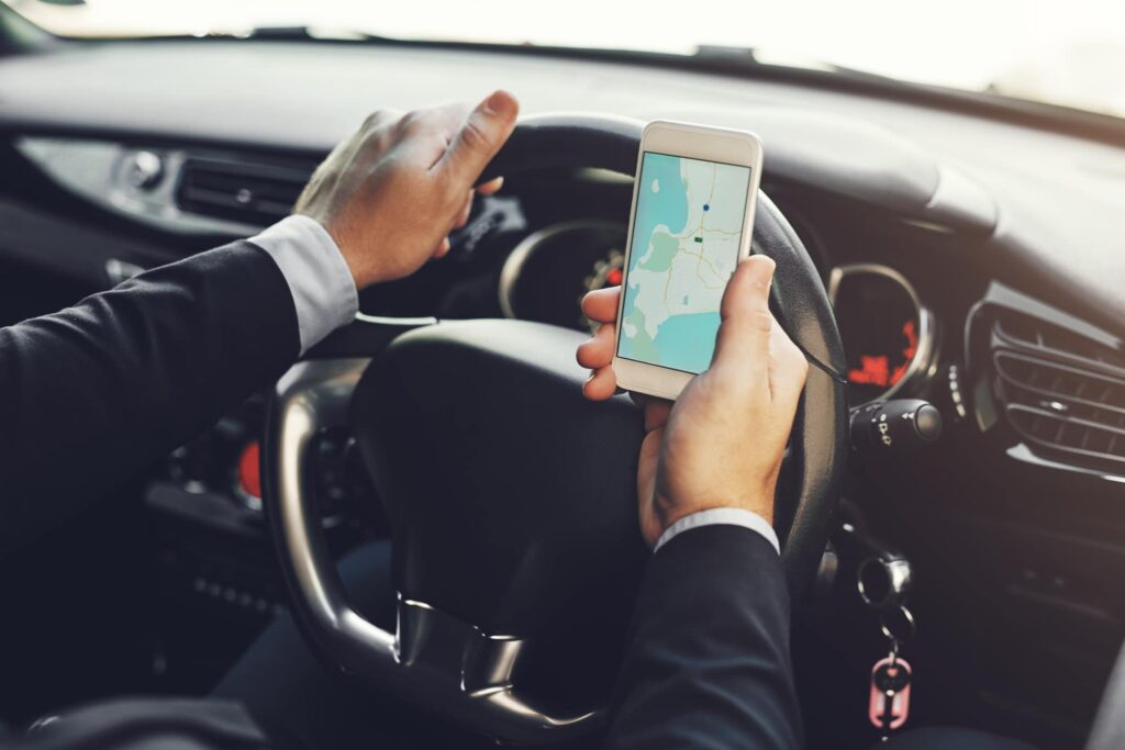 Person in a suit driving a car while holding a smartphone displaying a map with navigation directions.