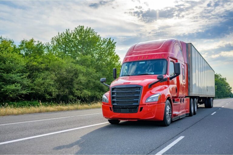 A red semi-truck with a large trailer drives on a highway bordered by trees under a partly cloudy sky.