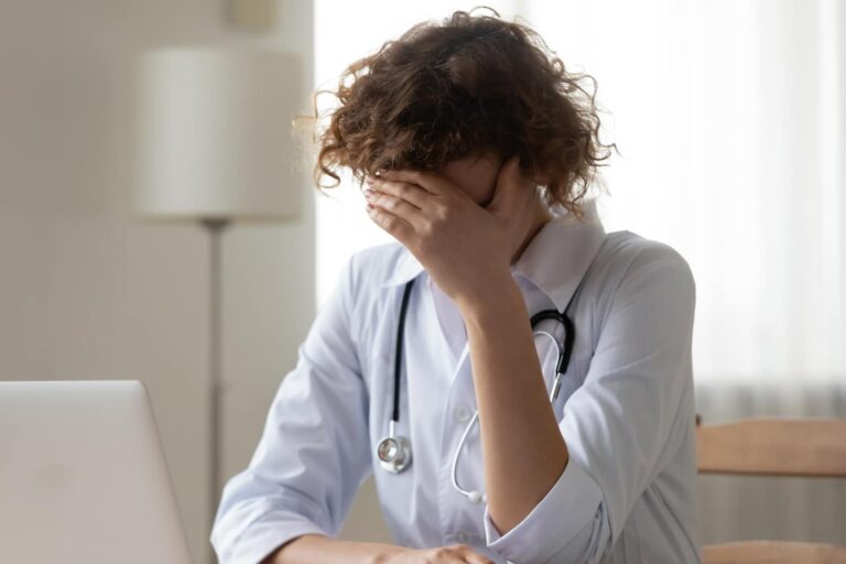 A person in a white lab coat with a stethoscope sits at a desk, covering their face with one hand, appearing stressed or tired.