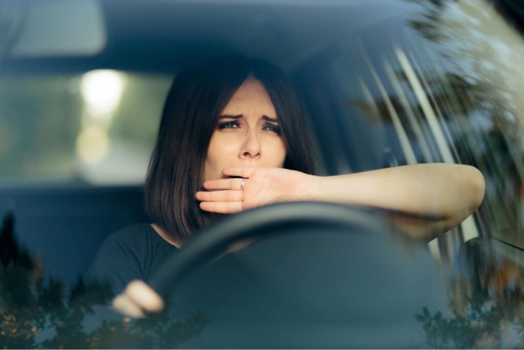 A woman sits in the driver's seat of a car, covering her mouth with her hand and appearing tired or sleepy.