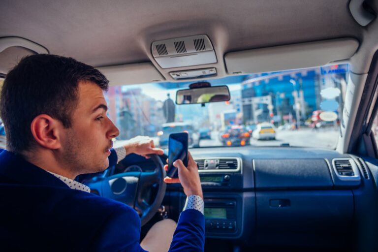 A man in a car holds a smartphone in his right hand while driving through city traffic on a sunny day.