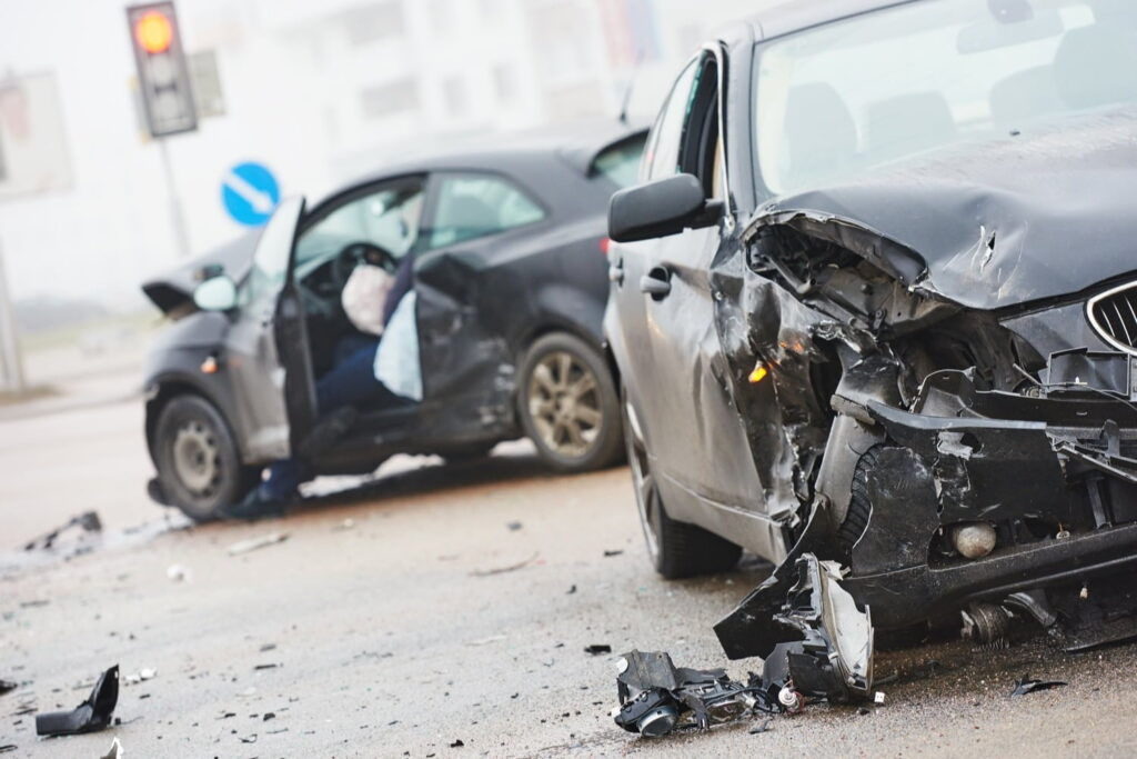 Two cars with significant front-end damage sit at an intersection after a collision. Both driver-side airbags have deployed, and debris is scattered on the road.
