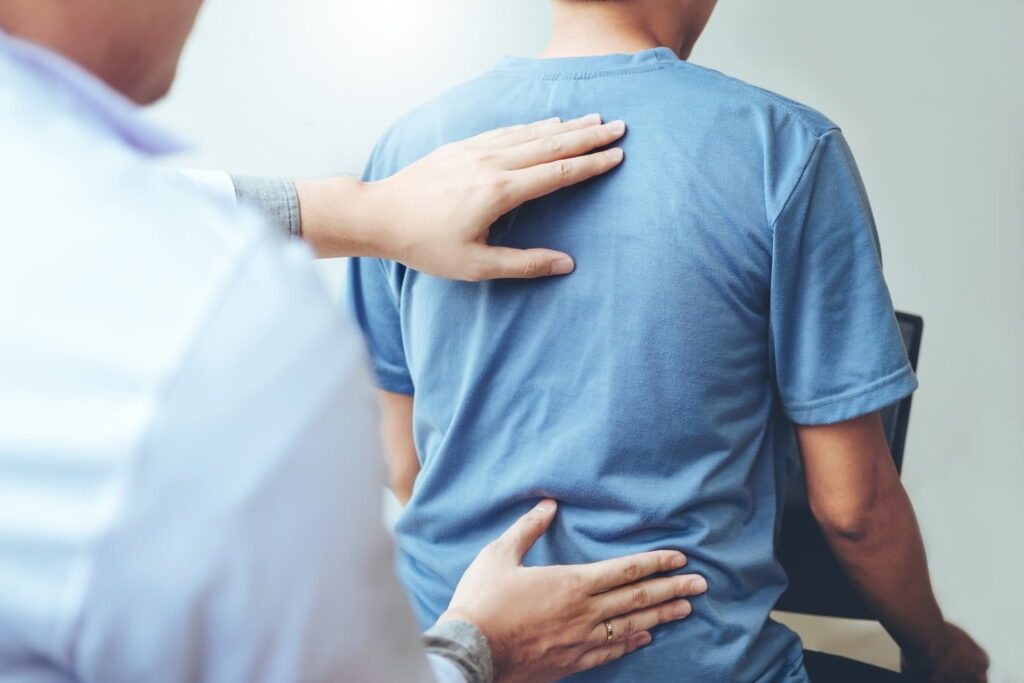 A healthcare professional examines a person's back by placing hands on the upper and lower back while the person sits upright.