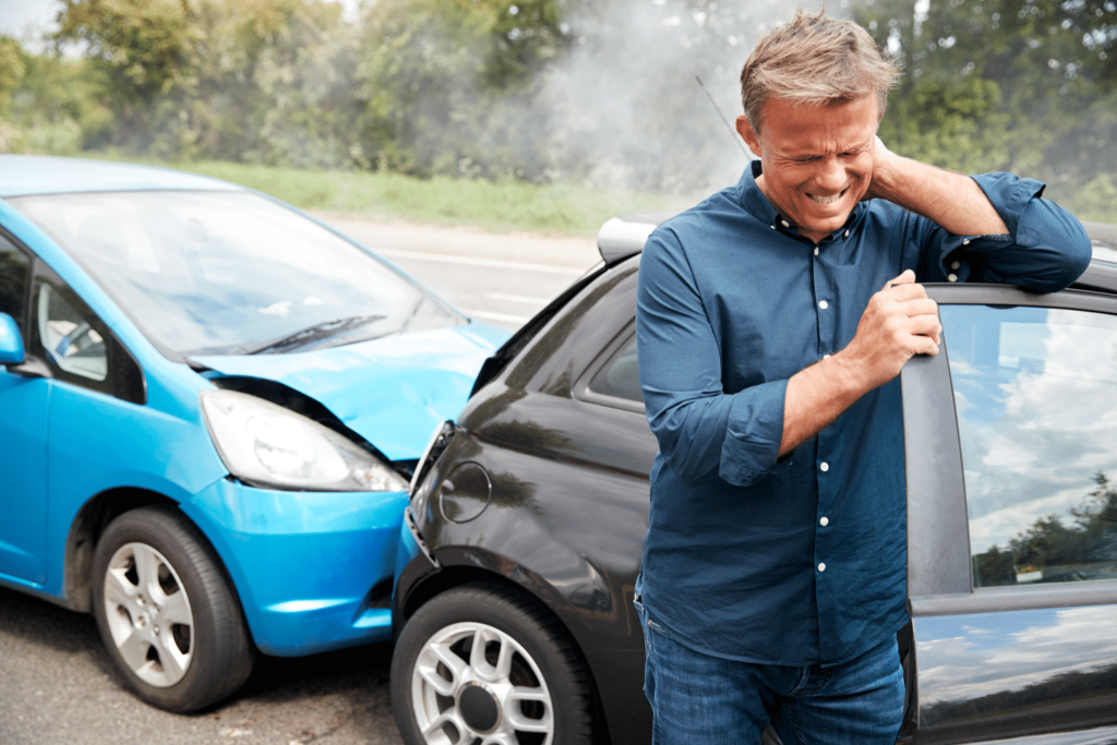 A man stands beside a black car holding his neck, appearing in pain, after a rear-end collision with a blue car on a road.