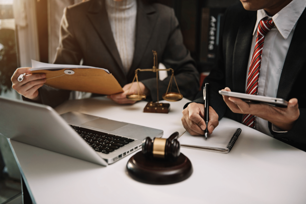 Two people in business attire sit at a desk with a laptop, gavel, legal scales, notebook, and documents, suggesting a legal or business meeting.