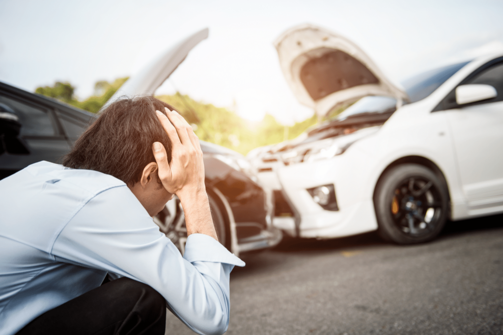 A person sits on the ground holding their head in their hands near two cars with open hoods after a collision.