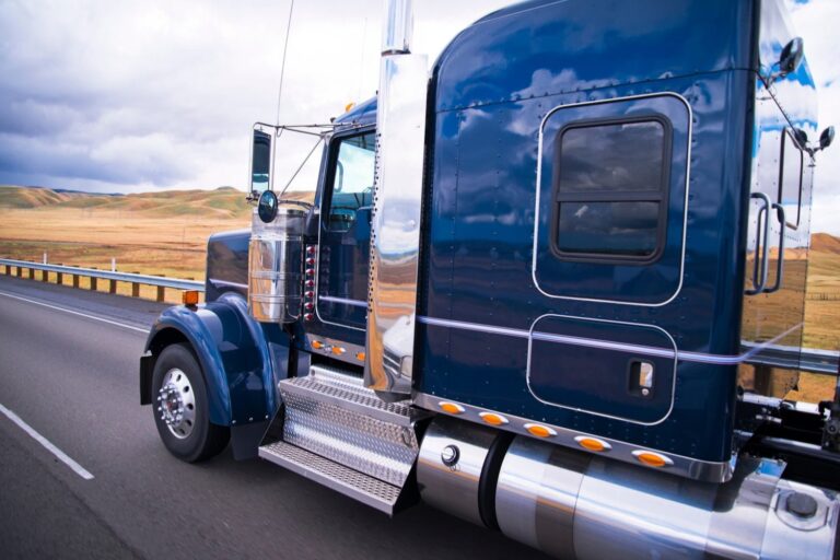 A blue semi-truck drives on a highway next to a metal guardrail, with open fields and cloudy skies in the background.