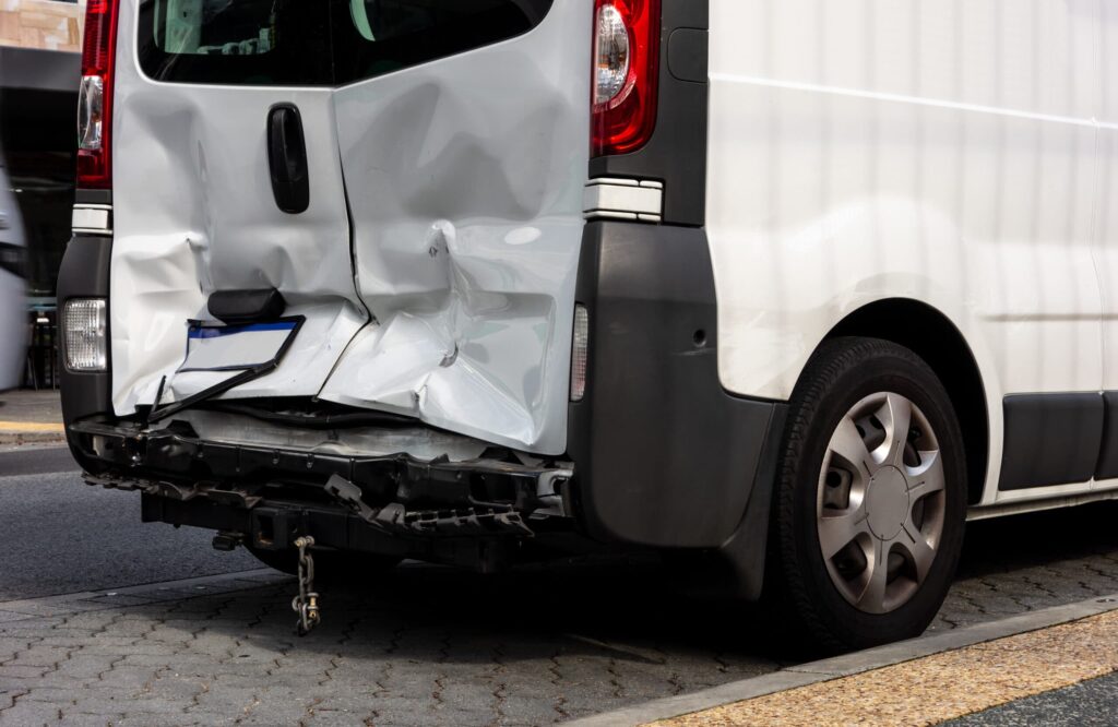 A white van with significant damage to its rear bumper and door is parked on the side of a street.