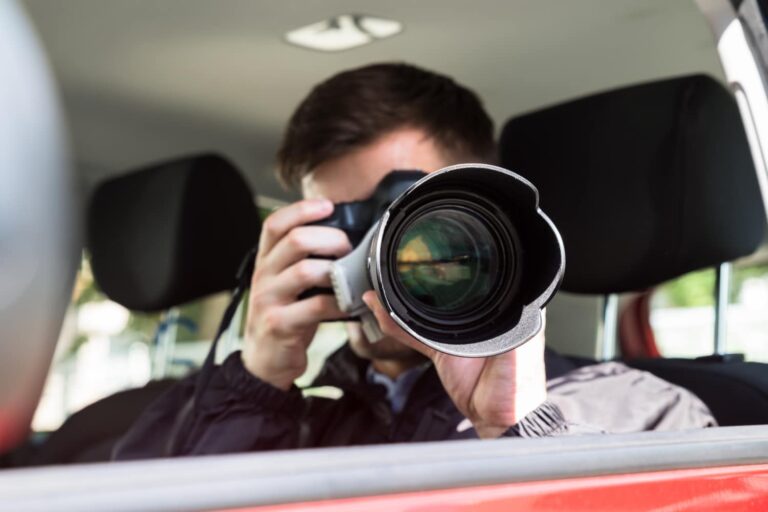 A person sitting in a car aims a camera with a large zoom lens out the window, taking a photograph.