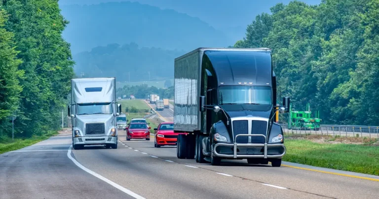 Several semi-trucks and cars drive on a multi-lane highway surrounded by trees and hills under a clear sky.