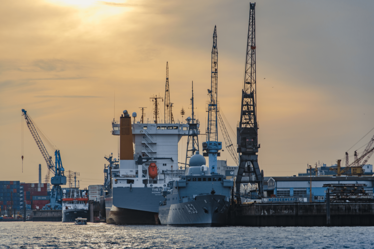 Several ships docked at an industrial port, surrounded by large cranes and warehouses under a cloudy sky during sunset.