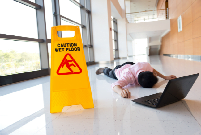A person lies on the floor near a laptop in a hallway, with a "Caution Wet Floor" sign placed nearby.