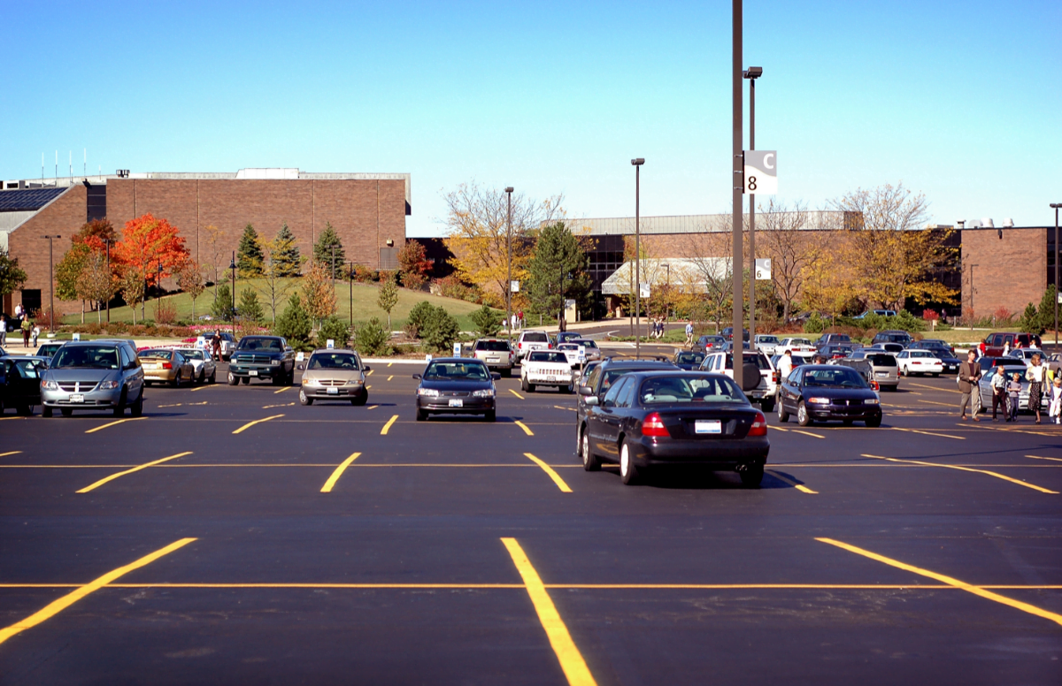 A large parking lot with several parked cars and a few empty spaces in front of a brick building and trees with autumn foliage under a clear blue sky.