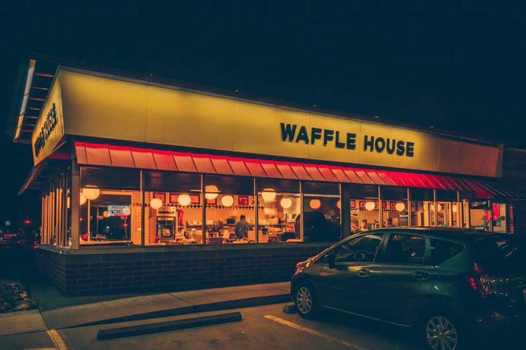 A Waffle House restaurant at night, brightly lit with a yellow sign, large windows showing diners inside, and a parked car in front.