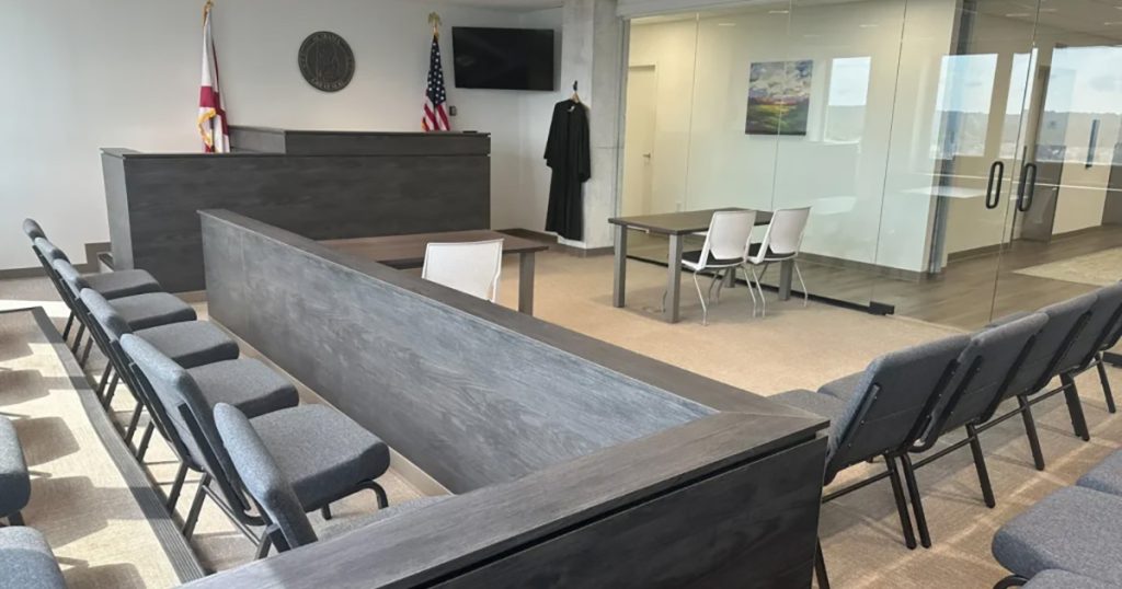 Modern courtroom with rows of chairs, a judge's bench, flags, a wall-mounted TV, and a table with white chairs; glass wall in the background.