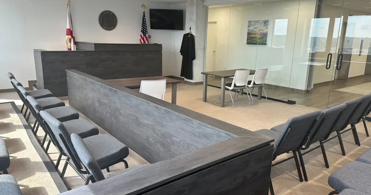 Modern courtroom with rows of chairs, a judge's bench, flags, a wall-mounted TV, and a table with white chairs; glass wall in the background.